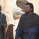 Obi-Wan Kenobi, wearing a poncho and blue robes, walks through a town on Tatooine.